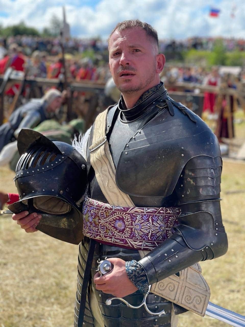 Portrait of a man wearing black plate armour and holding a helmet